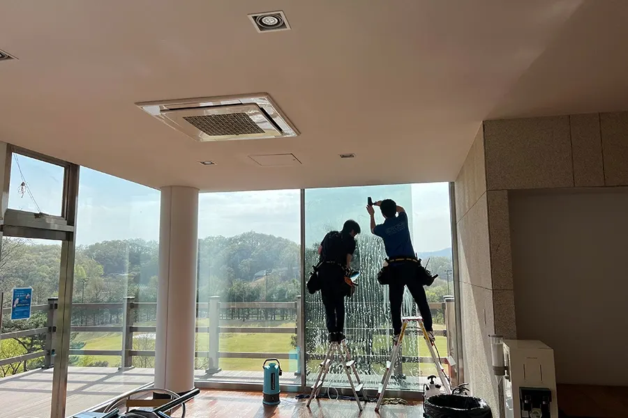 통유리 휴게 공간 창가에서 사다리를 놓고 유리열차단필름을 붙이는 시공 기사 두 명