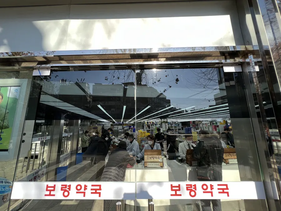 통유리 출입문 너머로 약국 내부와 손님들이 보이는 전경, 아침 햇살이 비치는 모습